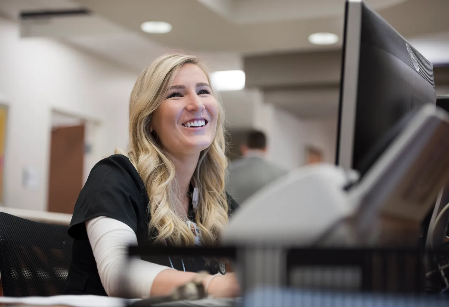 A Novant Health team member is sitting at a front desk smiling. A Novant Health team member is sitting at a front desk smiling.